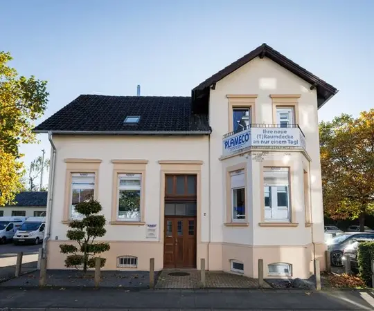 Helles Firmengebäude mit Plameco-Werbeschild für Spanndecken, umgeben von Bäumen und Autos.