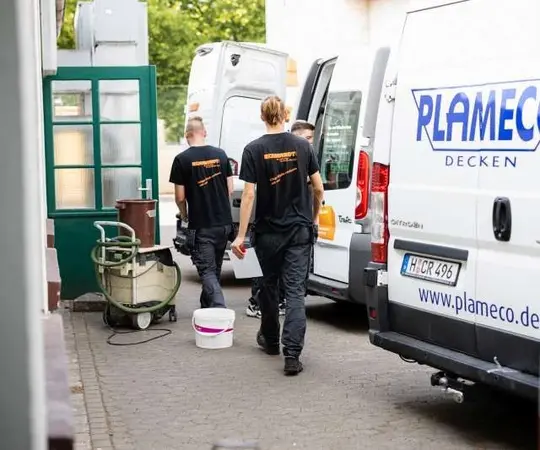 Handwerker tragen Arbeitsmaterial zu weißen Plameco Transportern vor einem Gebäude.