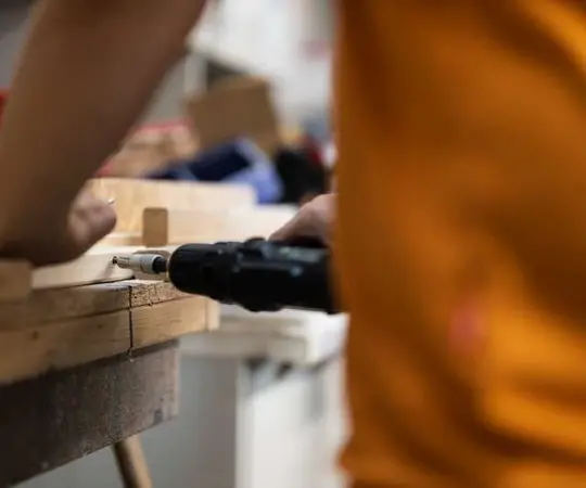 Eine Person schraubt mit einem Akkuschrauber Holz in einer Werkstatt zusammen.