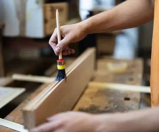 Handwerker trägt Holzschutzmittel mit Pinsel auf Holzbrett in einer Werkstatt auf.