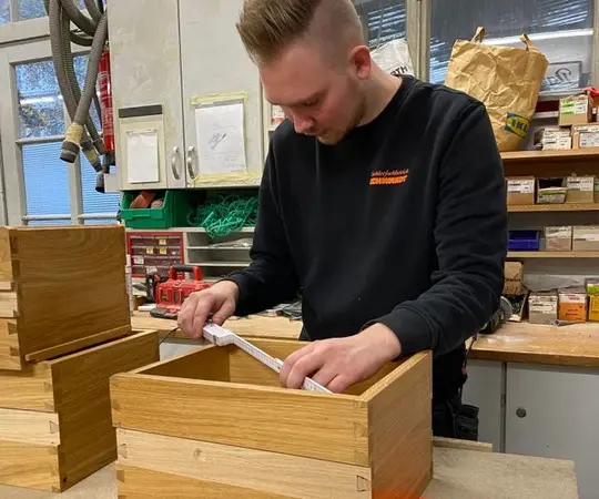Ein junger Mann misst in einer Werkstatt eine Holzkiste mit einem Zollstock aus.
